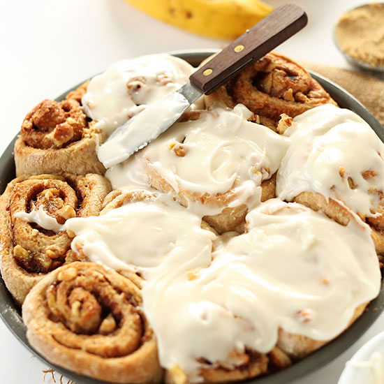 Spreader knife coated in frosting on a pan of Banana Bread Cinnamon Rolls