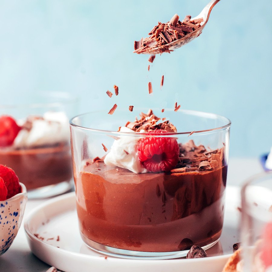 Sprinkling shaved dark chocolate onto a dairy free pot de creme topped with coconut whipped cream and a fresh raspberry