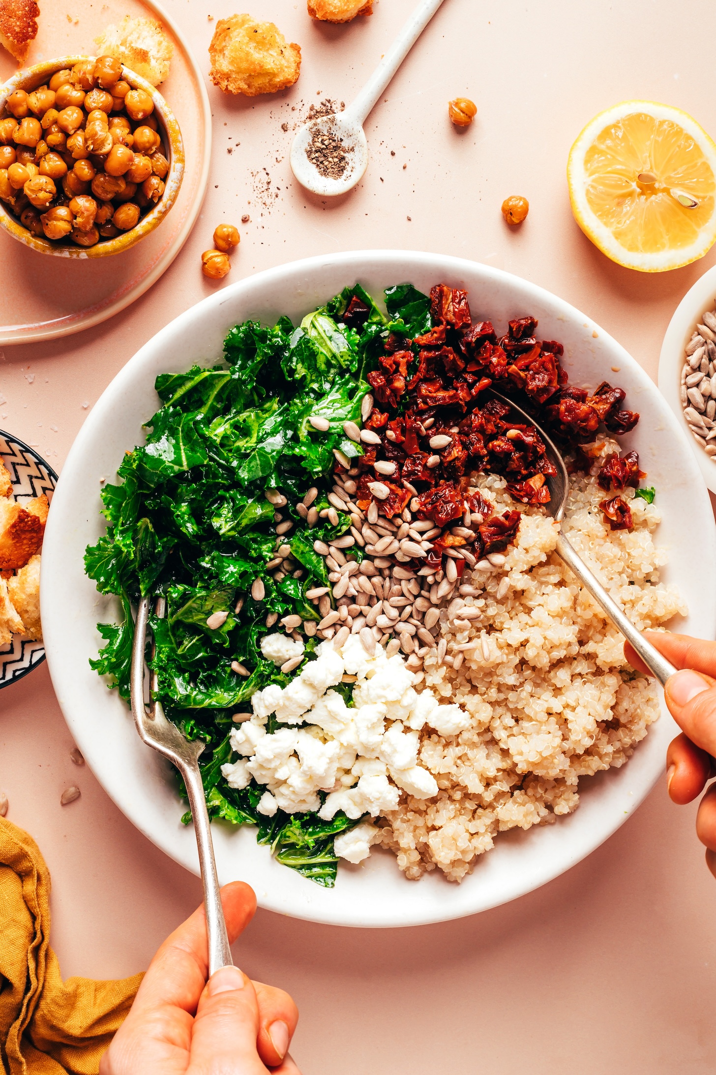 Using a fork and spoon to toss a bowl of sun-dried tomatoes, quinoa, sunflower seeds, goat cheese, and massaged kale