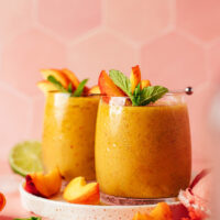 Two glasses of our frozen peach daiquiri with sliced peaches and mint resting at the top of the glasses