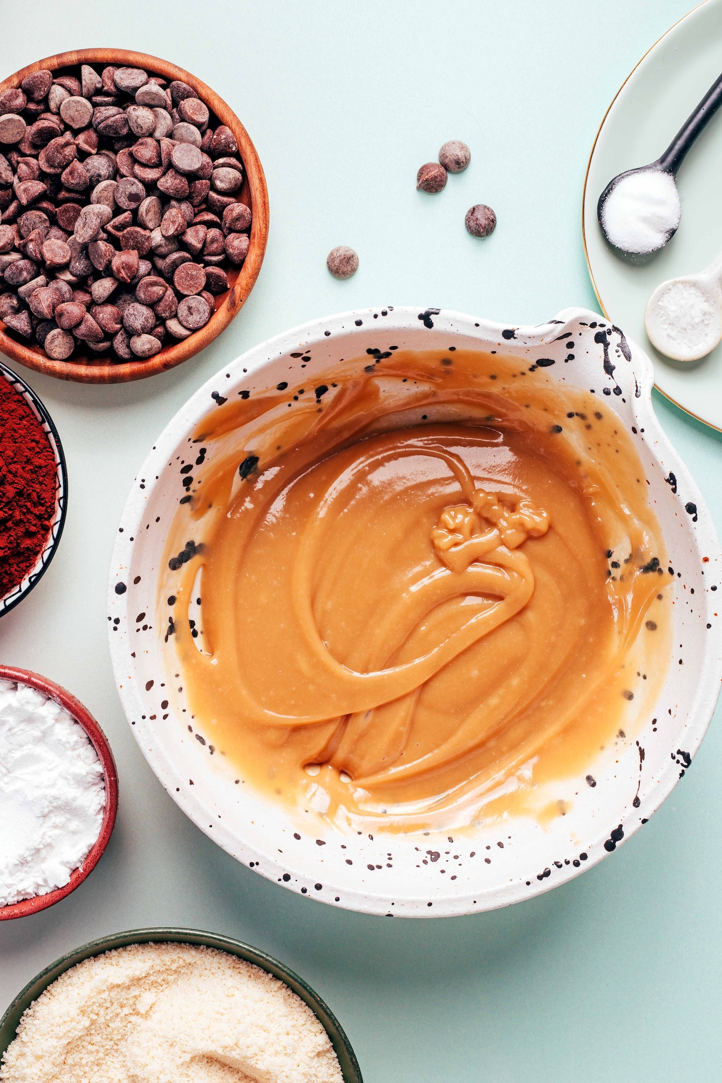 Bowl of creamy whisked tahini and maple syrup next to salt, baking soda, chocolate chips, cocoa powder, tapioca starch, and almond flour