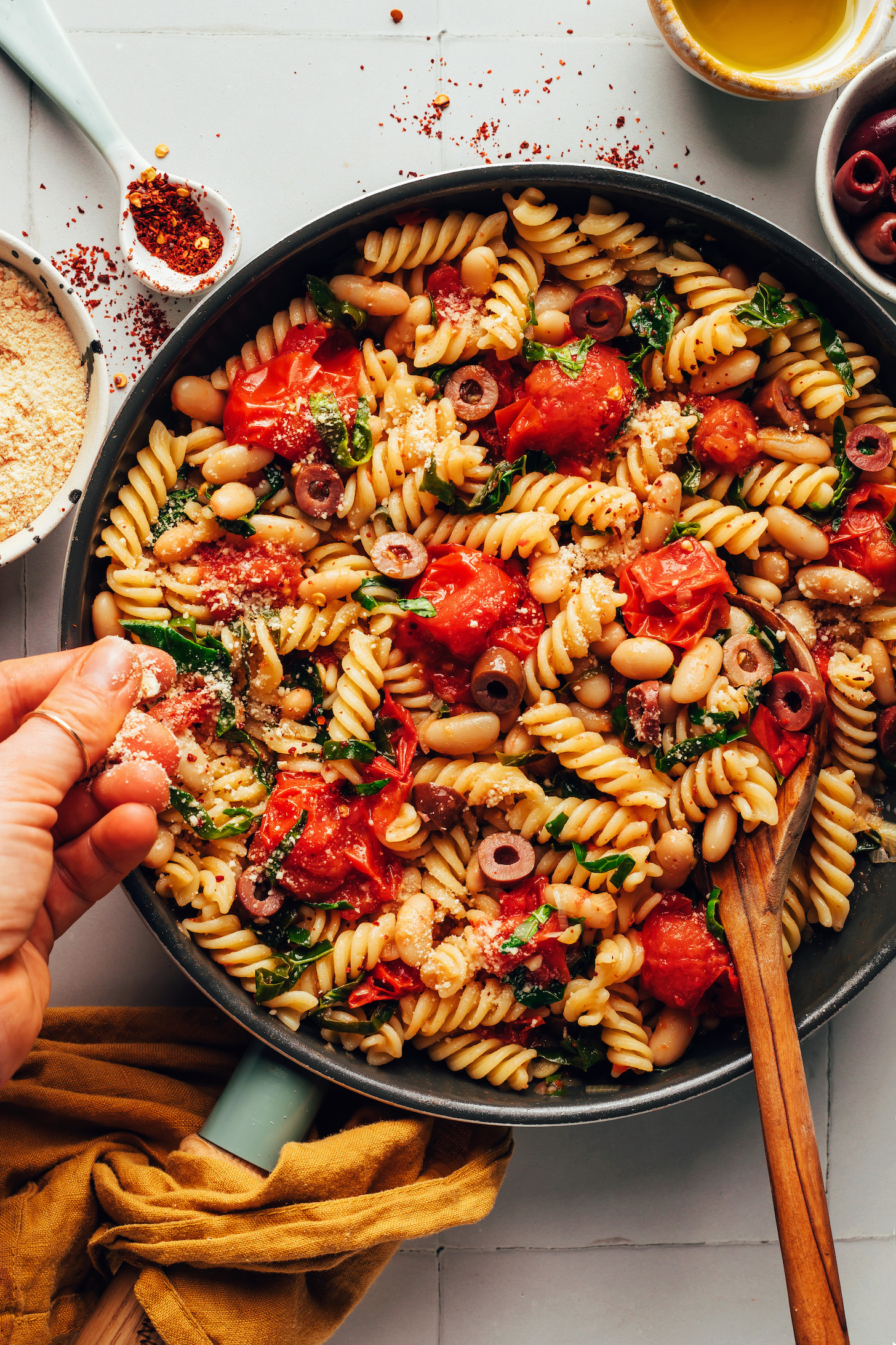 Sprinkling vegan parmesan cheese over a pan of garlicky cherry tomato and white bean pasta