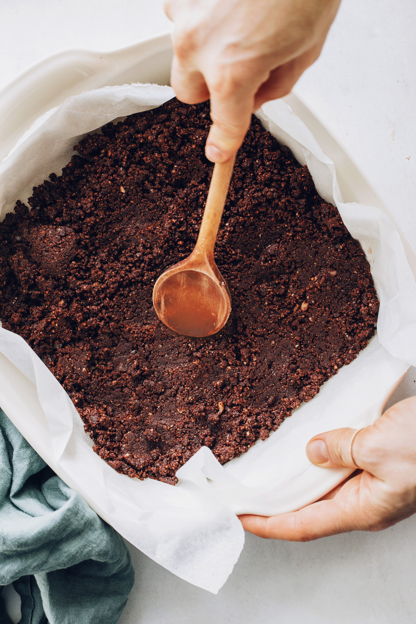 Using a wooden spoon to spread the chocolate brownie layer in a parchment-lined pan
