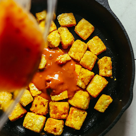 Pouring peanut sauce into a skillet of Crispy Peanut Tofu