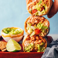 Holding a vegan crunchwrap-inspired burrito above a stack of more burritos