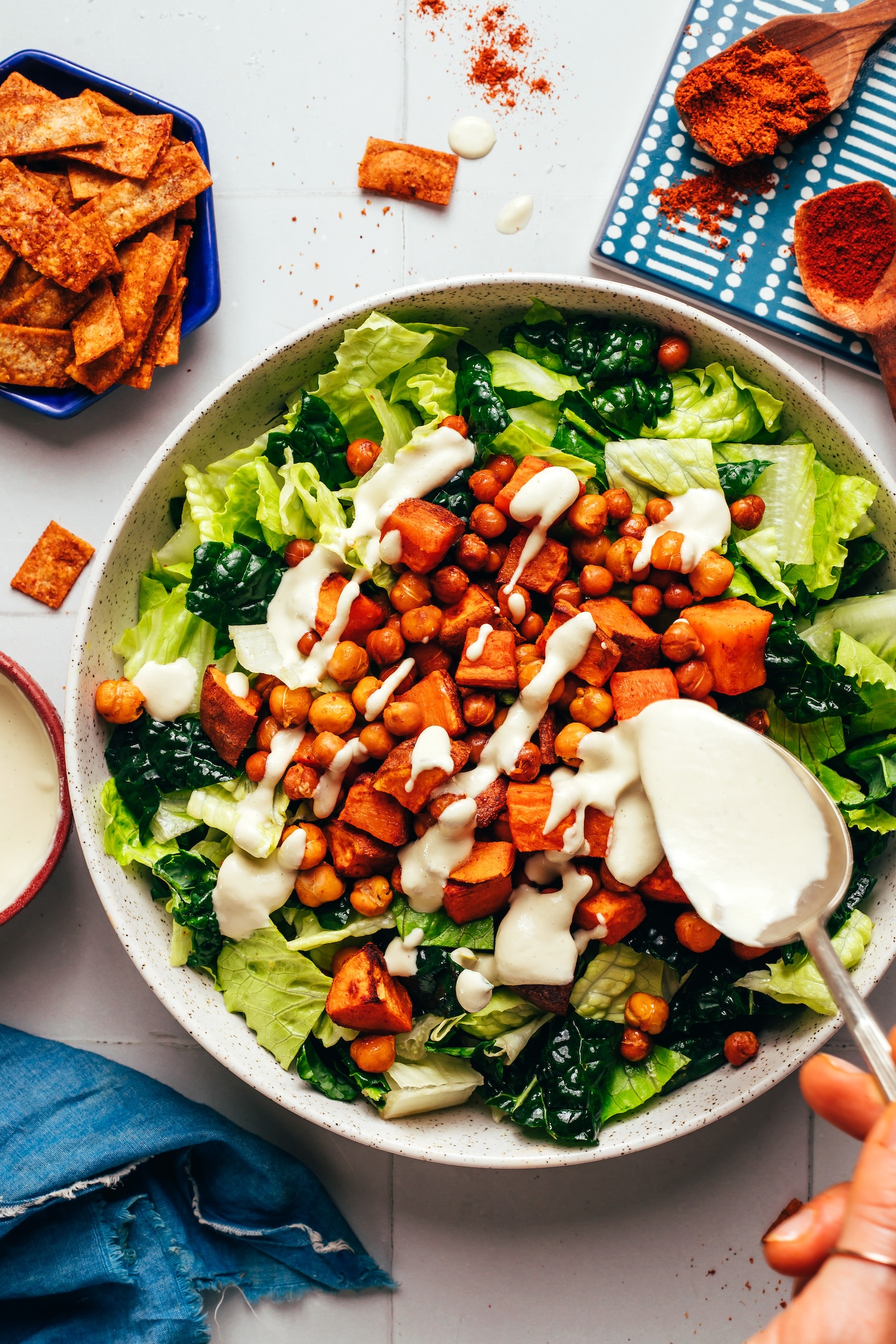 Using a spoon to pour vegan cashew jalapeño dressing onto greens and roasted sweet potato and chickpeas