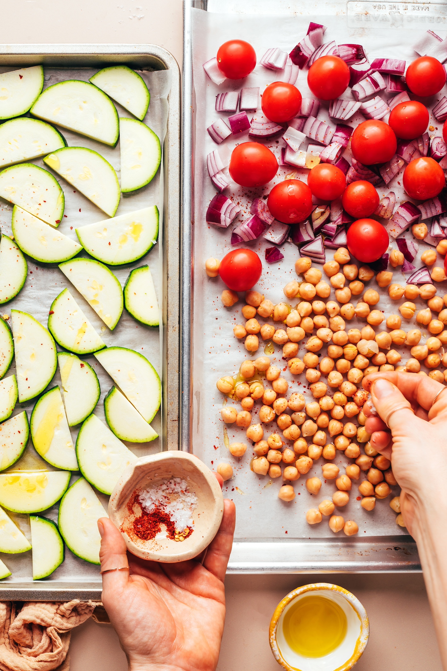 Sprinkling spices onto chickpeas on a baking sheet with tomatoes, red onion, and zucchini
