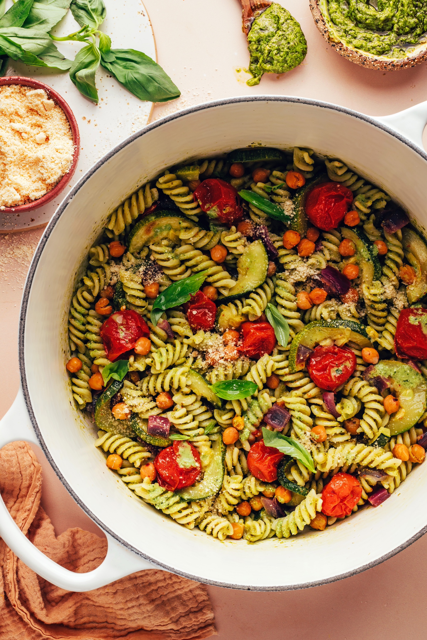 Bowl of vegan parmesan next to a Dutch oven filled with Zucchini Pesto Pasta with Roasted Tomatoes & Chickpeas